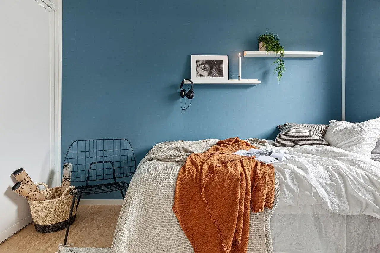 Camera da letto con pareti blu intenso, un letto accogliente con coperte arancioni e bianche, e una sedia in metallo nero. Ottime idee pareti bicolore per un ambiente moderno.