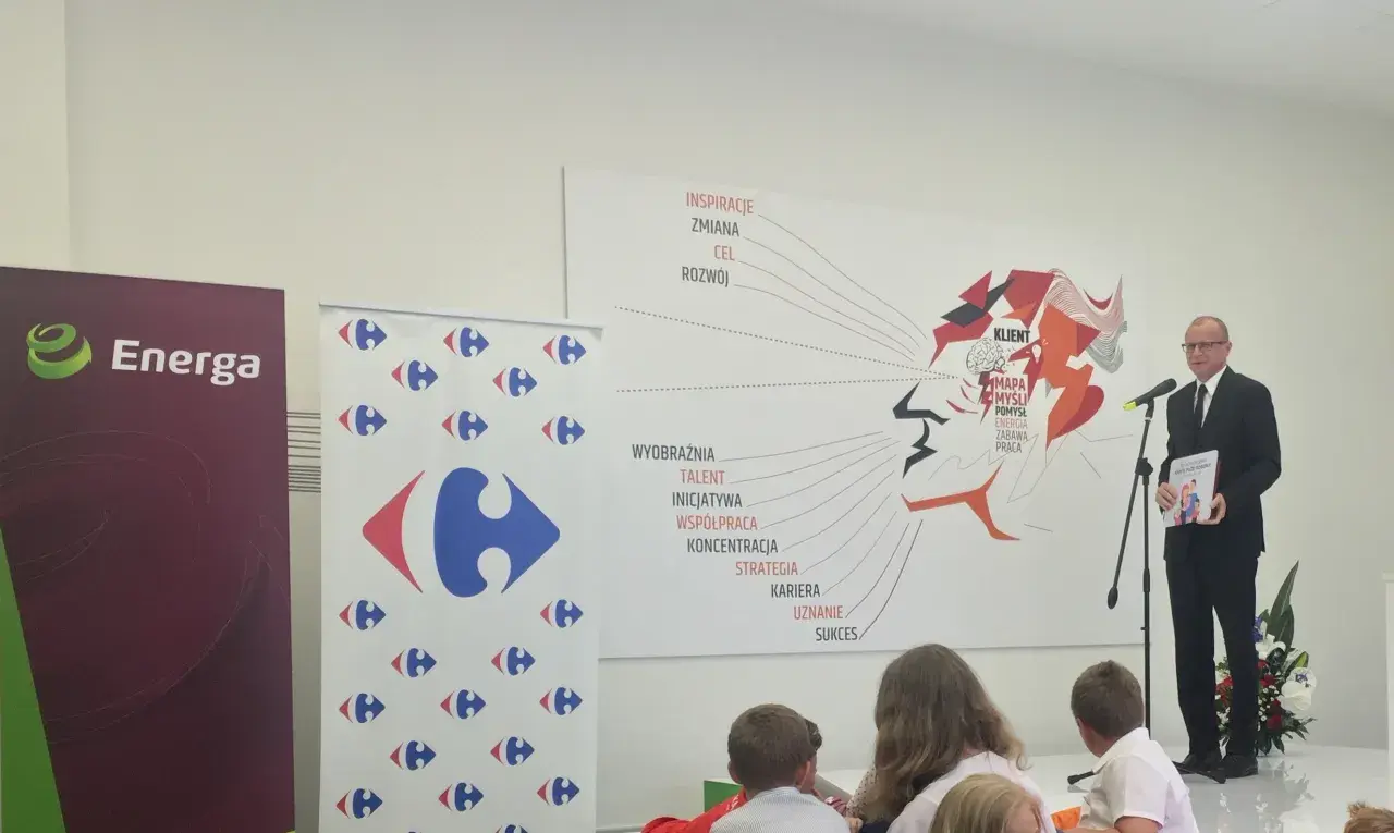 Mężczyzna w garniturze wręcza książkę "Karta Dużej Rodziny" na tle banerów Energa i Carrefour.