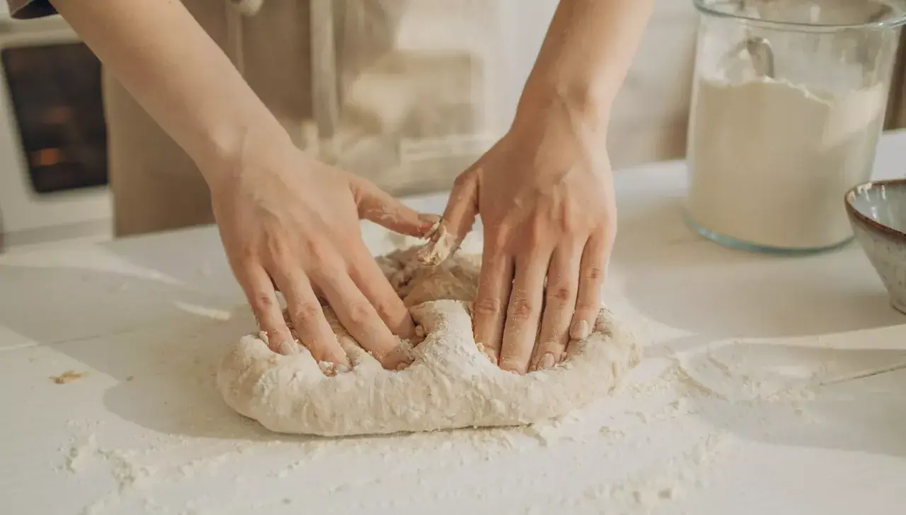 Manos amasando masa pan sobre una mesa con harina esparcida. Un frasco de vidrio con harina y un cuenco están al fondo.