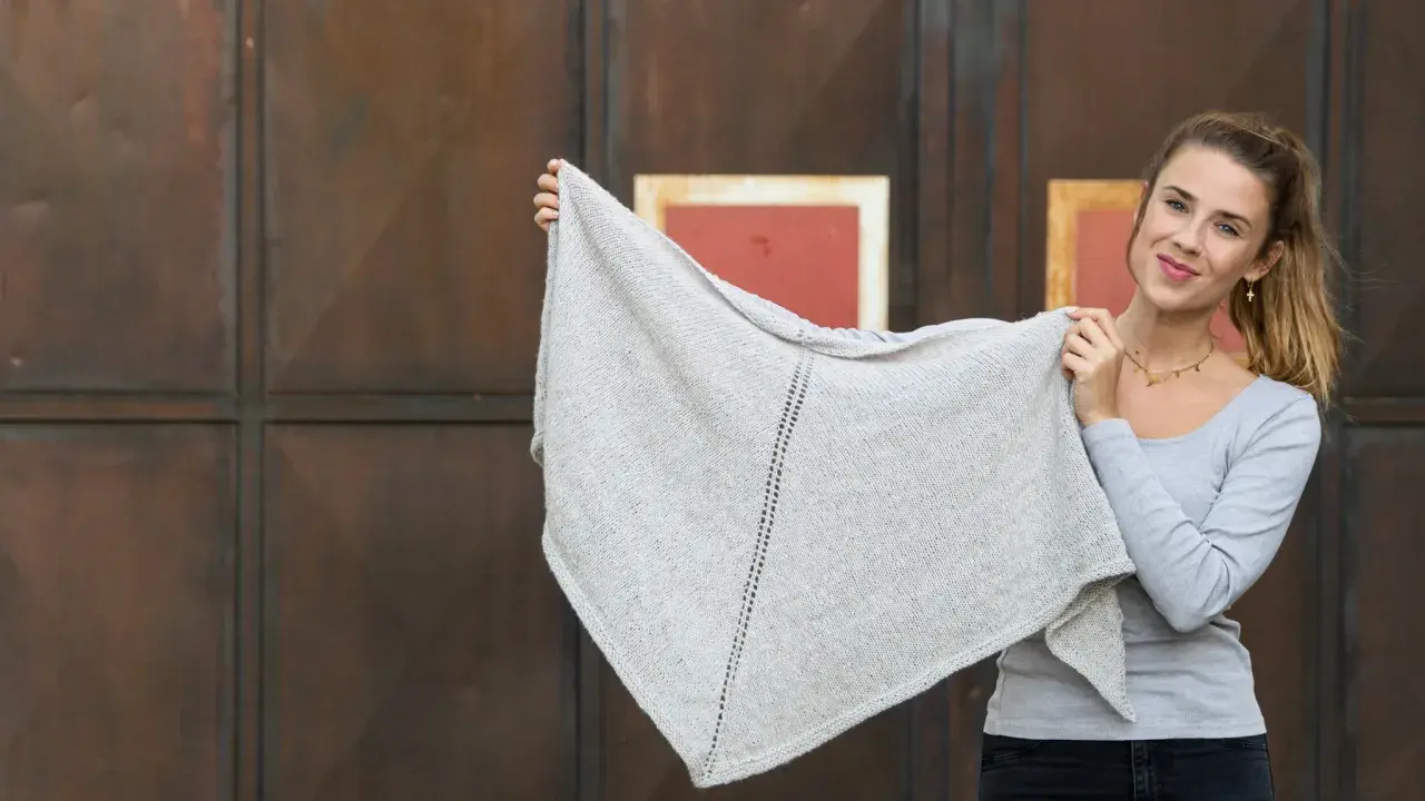 Junge Frau hält ein hellgraues, gestricktes Dreieckstuch hoch.