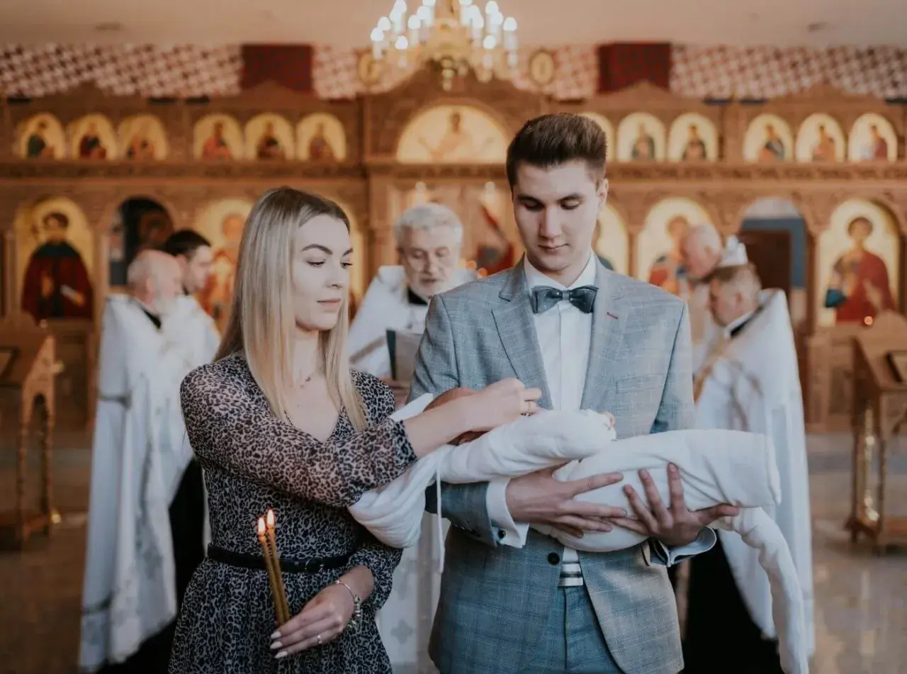 Młodzi rodzice trzymają dziecko do chrztu prawosławnego. W tle widać cerkiew i duchownych.