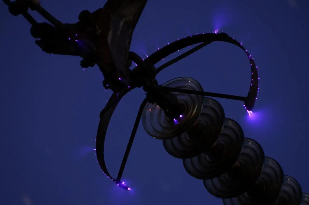Des arcs électriques violets, les feux de Saint-Elme, crépitent sur une structure métallique et des isolateurs en verre contre un ciel nocturne.