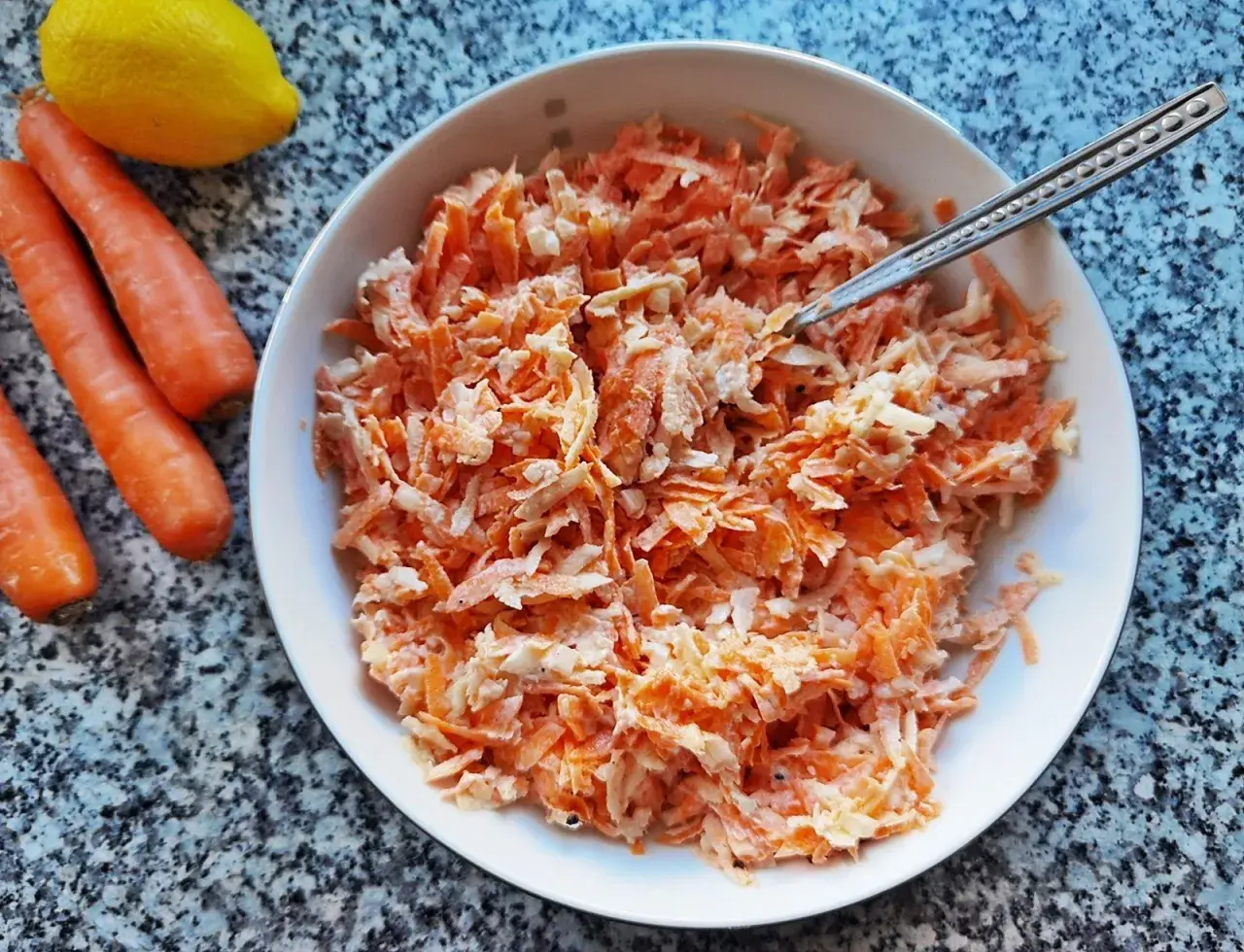 Pyszna surówka z marchewki jak z restauracji, gotowa do podania. Obok leżą świeże marchewki i cytryna.