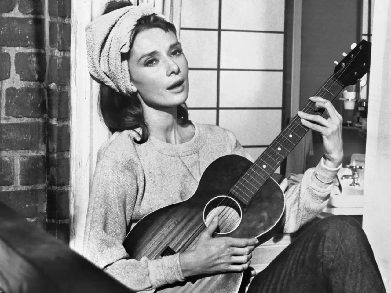 Audrey Hepburn, z ręcznikiem na głowie, gra na gitarze, śpiewając "Moon River".