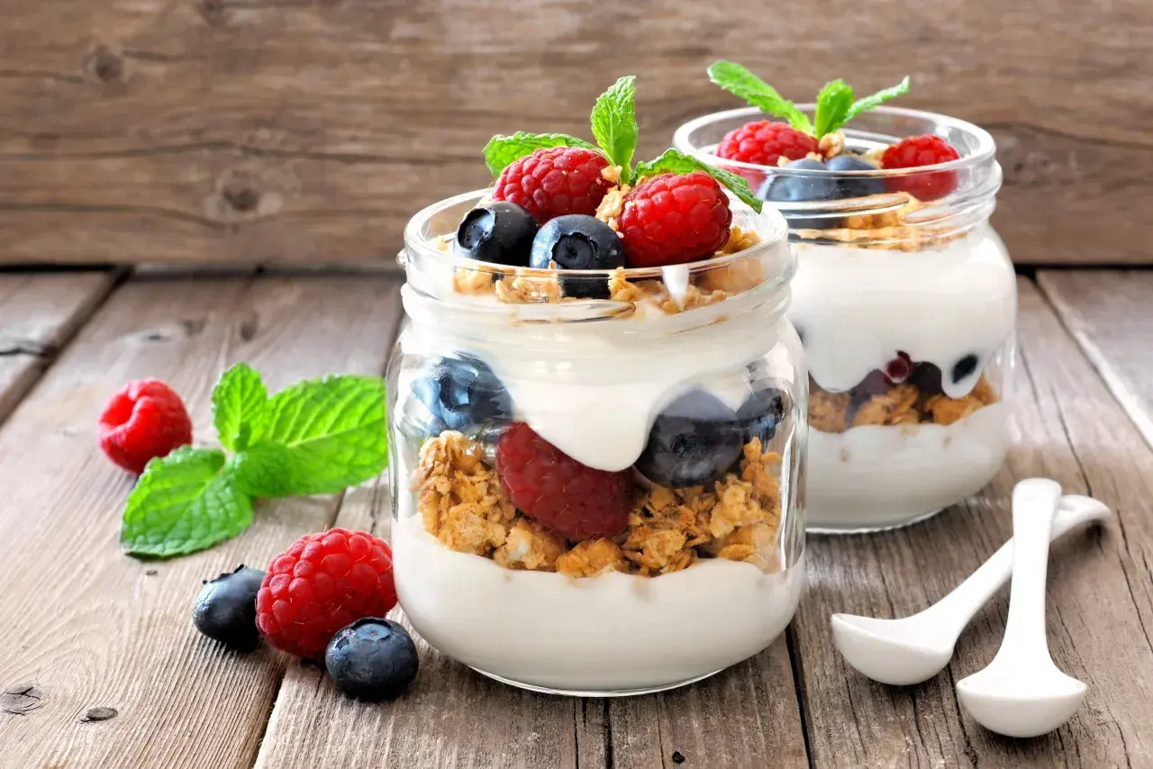 Dos frascos de vidrio con yogur, granola, frambuesas y arándanos. ¡Cuidado! El yogur con avena engorda si no se consume con moderación.