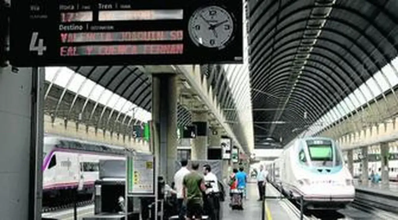 Un tren de alta velocidad espera en la estación. El panel de información muestra un destino a Valencia y Sevilla.