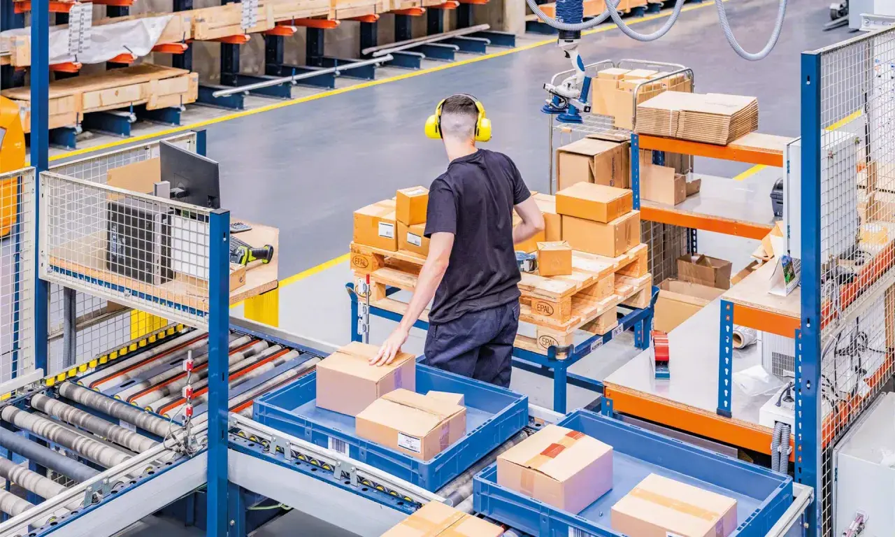 Ein Mitarbeiter mit Kopfhörern sortiert Pakete in einem Lager. Hier wird alles **assemble to order** für den Versand vorbereitet.