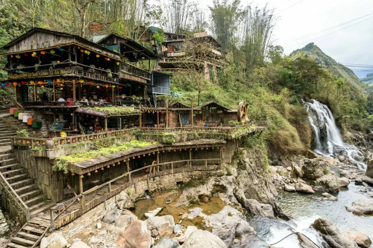 Holzgebäude schmiegen sich an einen Hang in Sapa, Vietnam, mit einem Wasserfall, der daneben herabstürzt.