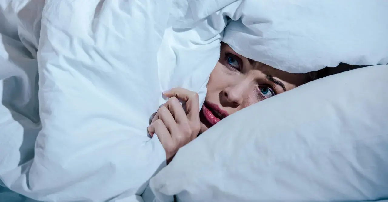 Verängstigte Frau versteckt sich unter der Decke, die Augen weit aufgerissen, als ob sie einen Albtraum erlebt.