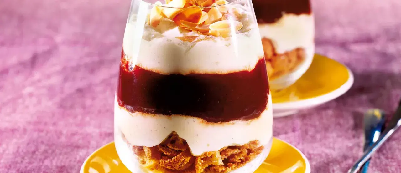 Schichtdessert im Glas mit zerbröselten Amarettini, roter Fruchtsauce und cremiger Masse, garniert mit Mandelblättchen.