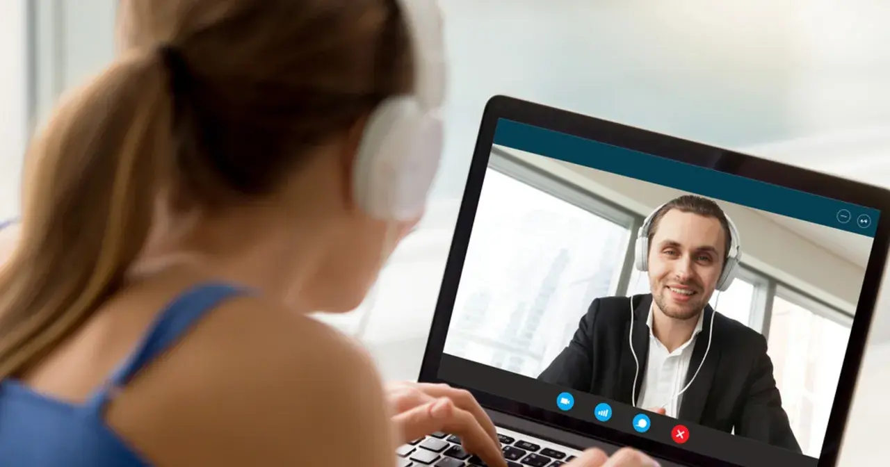 Kobieta w słuchawkach pisze na laptopie, prowadząc wideokonferencję z uśmiechniętym mężczyzną. On też ma słuchawki, ale ona pisze ale się nie umawia.