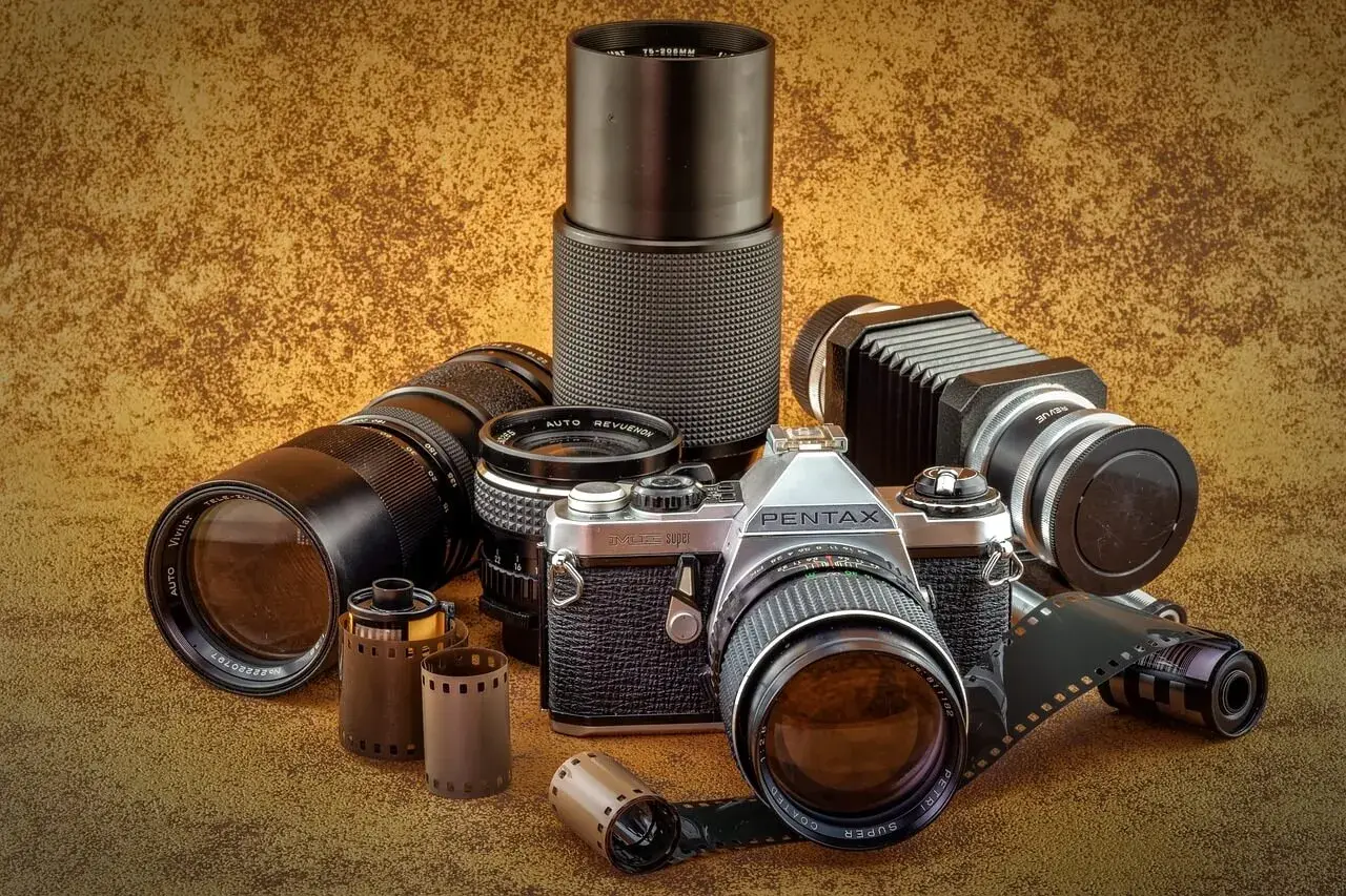 Kolekcja różnych rodzajów aparatów fotograficznych i akcesoriów: lustrzanka Pentax, obiektywy, mieszek, filmy.
