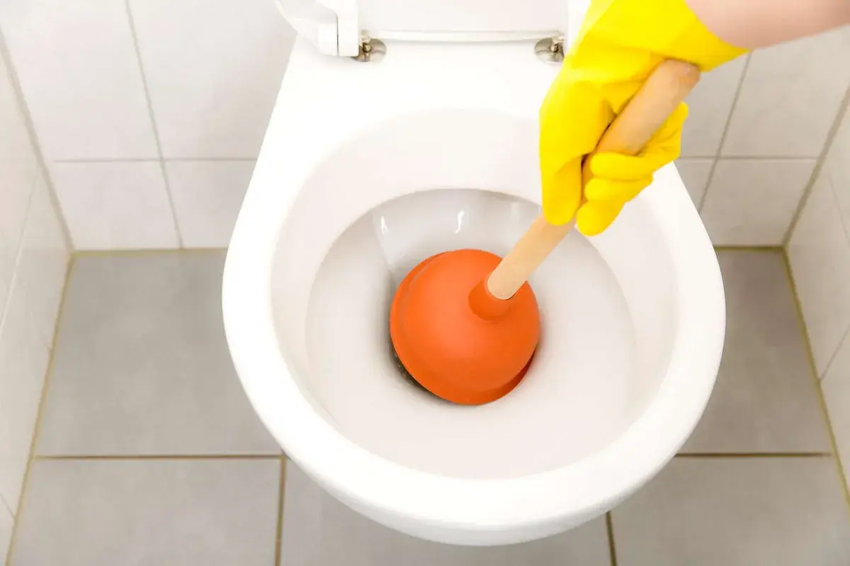 Mano con guanto giallo usa uno stantuffo arancione per liberare un filo d'acqua che scende nel water.