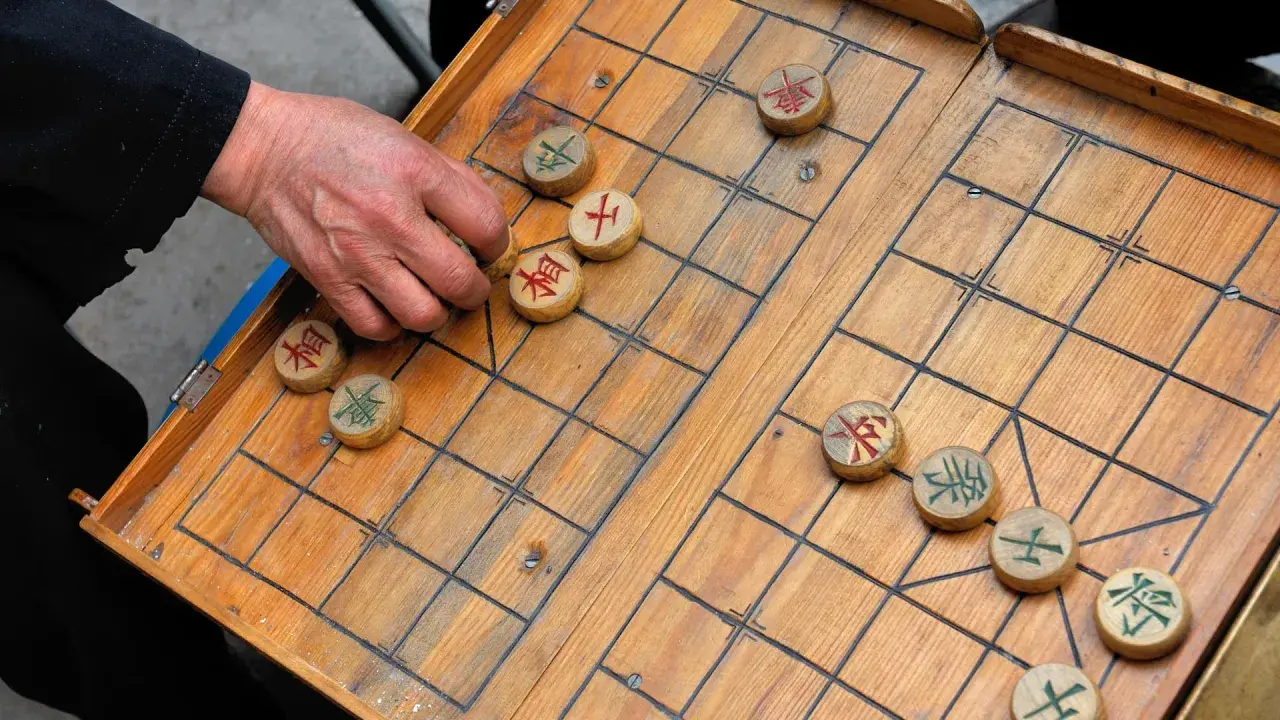 Eine Hand bewegt einen Stein beim China Schach. Das Holzbrett ist aufgeschlagen, die Figuren mit chinesischen Schriftzeichen sind verteilt.