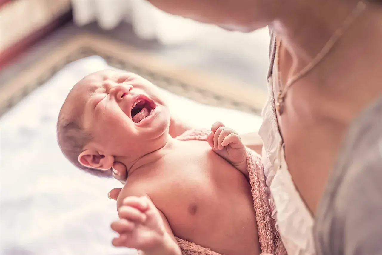 Bebé llorando con espasmos infantiles, envuelto en una manta rosa, sostenido por una persona.
