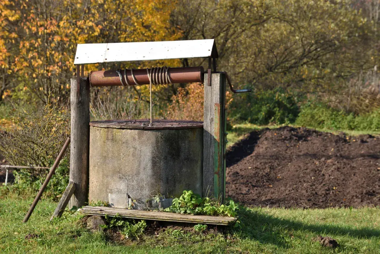 Stary, betonowy krąg studni kręgowej z metalową korbą i liną, otoczony jesienną zielenią.