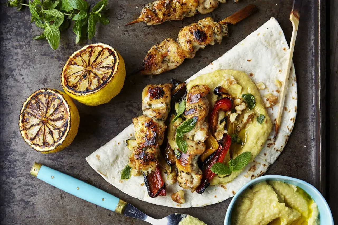 Grillad kyckling kebab med rostade grönsaker och hummus på ett tunnbröd, serverad med grillade citroner och mynta.