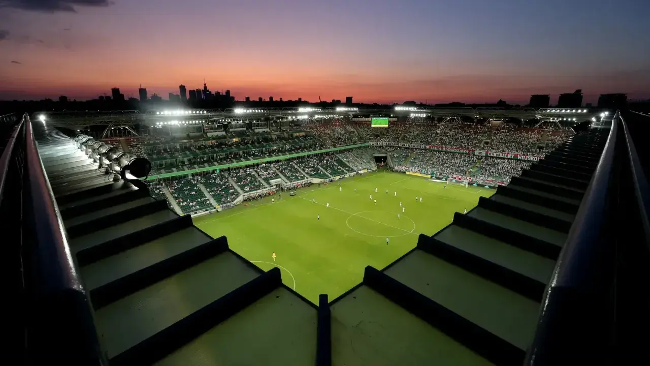 Zachód słońca nad stadionem Legii. Widok z góry na murawę, trybuny wypełnione kibicami i panoramę Warszawy. Pojemność stadionu Legii robi wrażenie.