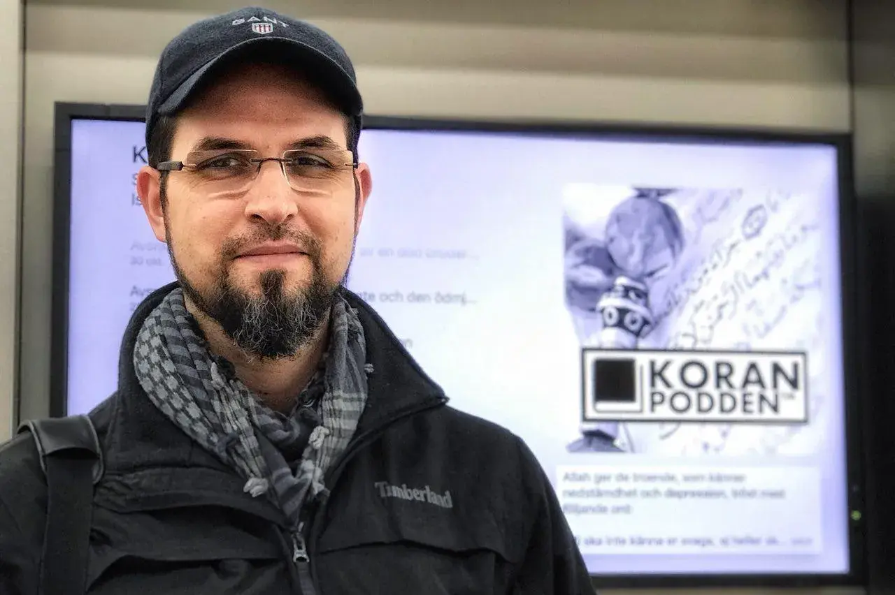 Man med skägg och keps framför en skärm med texten "KORAN PODDEN". Han ser tacksam ut, som om han ber: "ربي اوزعني ان اشكر نعمتك".