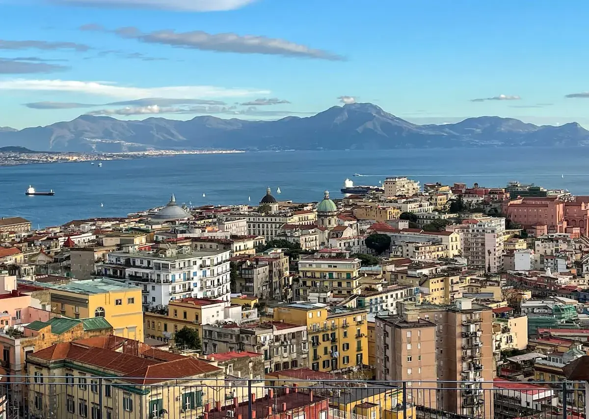 A panoramic view of Naples in December, with the blue sea and distant mountains under a cloudy sky.
