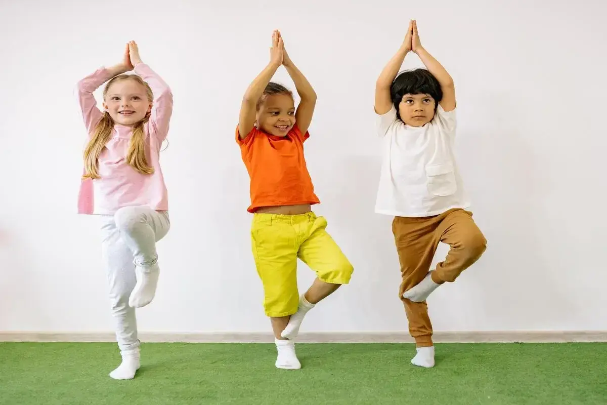 Tre bambini praticano yoga, in piedi su una gamba sola, con le mani giunte sopra la testa.