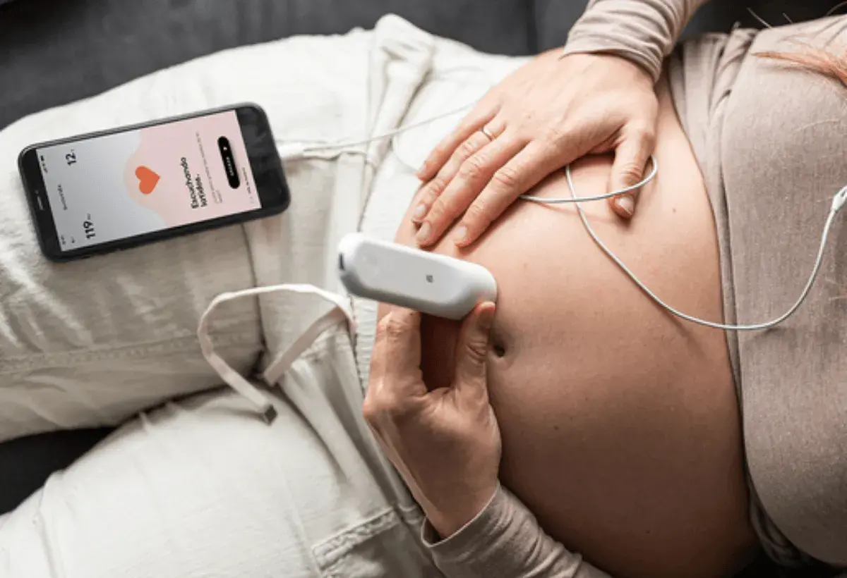 Manos de mujer embarazada usando un doppler fetal para escuchar el latido del bebé. La pantalla del móvil muestra "Escuchando latidos".