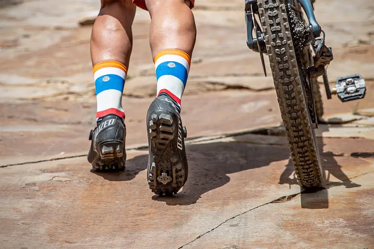 Mountain biking shoes with cleats are shown, not hiking boots. The rider's colorful socks and bike tire are also visible.