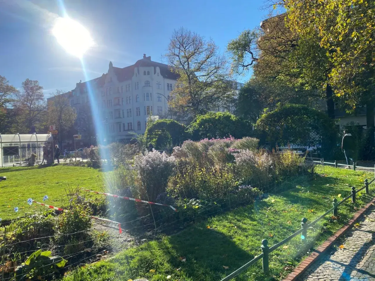 Grüner Park mit Blumenbeeten, Bäumen und einem Altbau unter strahlender Sonne.