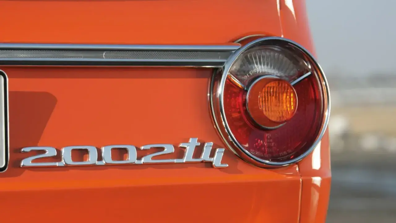 Detalle de la parte trasera de un BMW 2002 Tii naranja, con su icónico emblema cromado y una luz trasera circular.