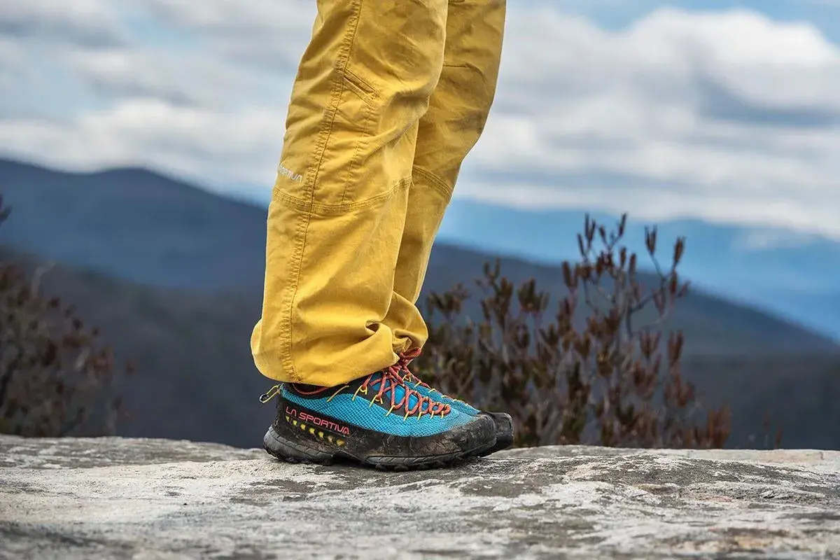 A person stands on a rock, wearing yellow pants and blue approach shoes with orange laces, ready for an adventure.