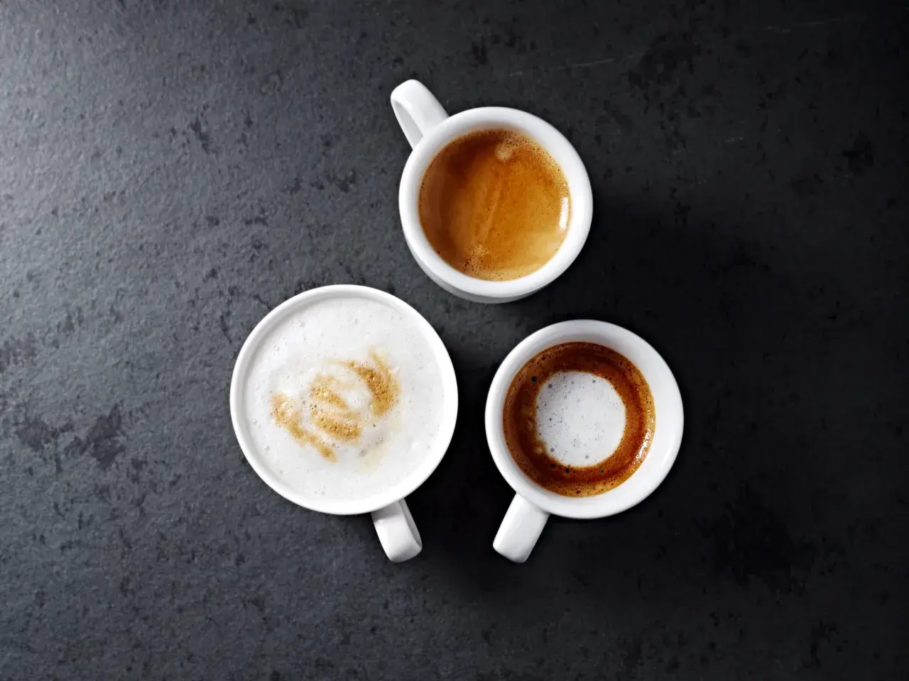 Trzy białe filiżanki z kawą na ciemnym tle. Jedna to espresso, druga to kawa macchiato z pianką, a trzecia to latte.