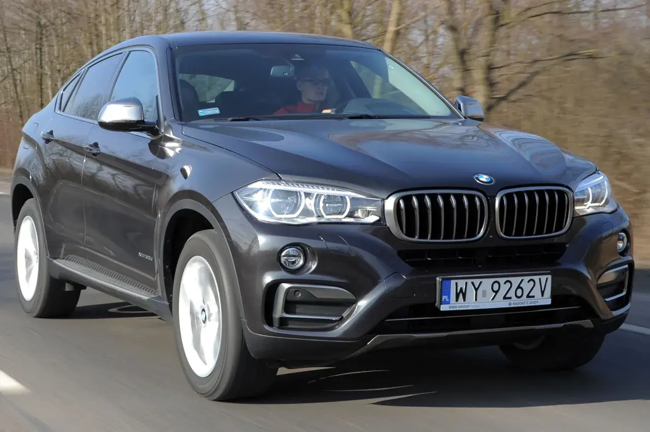 Ciemnoszary BMW F16 z kierowcą w czerwonej bluzie jedzie po drodze.