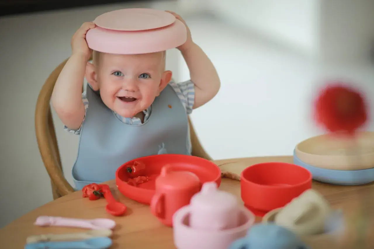 A smiling baby with a pink plate on its head sits at a table with baby feeding dishes.