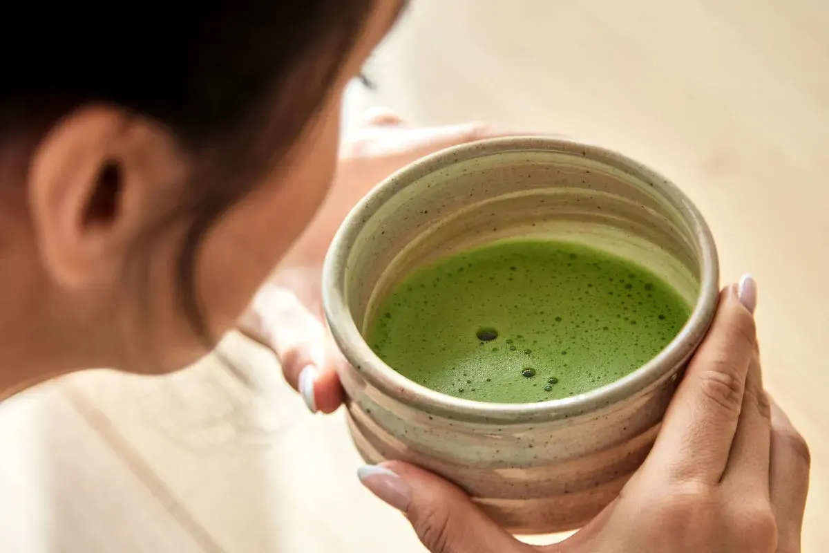 Frau genießt die Matcha Zubereitung in einer traditionellen Schale. Das leuchtend grüne Getränk schäumt leicht.