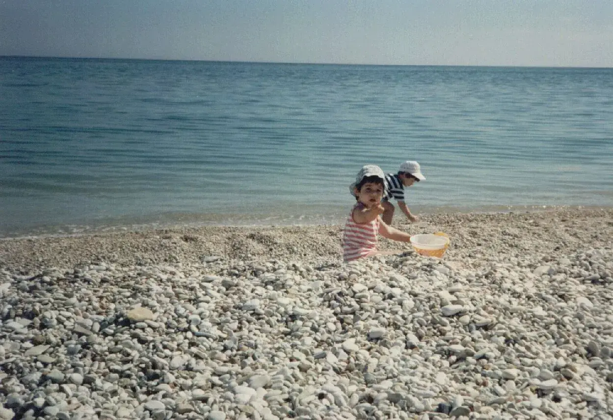 Bambini giocano sulla spiaggia di ciottoli a Marina di Montemarciano, con il mare azzurro sullo sfondo.