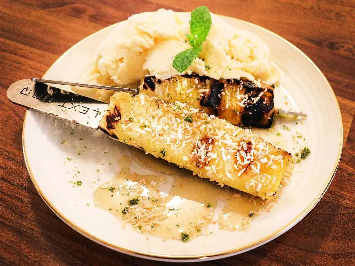 Grillad ananas på spett, toppad med kokosflingor, serveras med vaniljglass och en myntakvist. En härlig dessert!