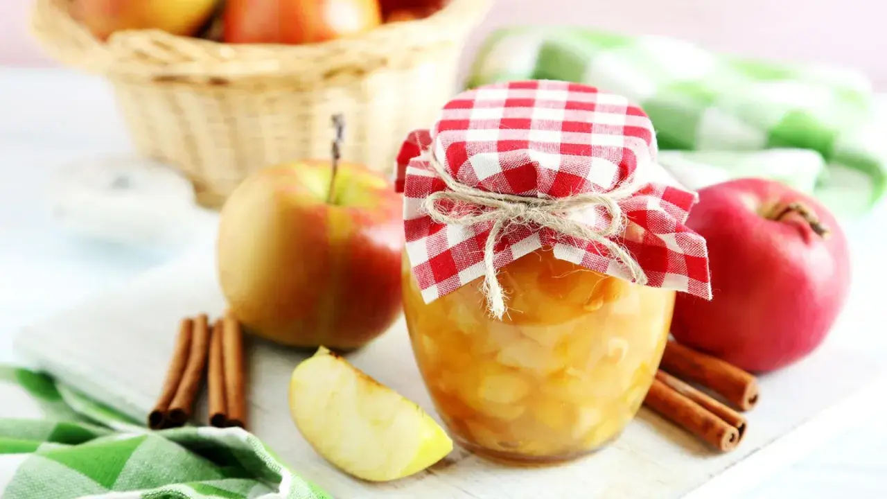Frisch eingekochtes Apfelkompott im Glas, dekoriert mit kariertem Stoff und Zimtstangen. Äpfel im Korb im Hintergrund.
