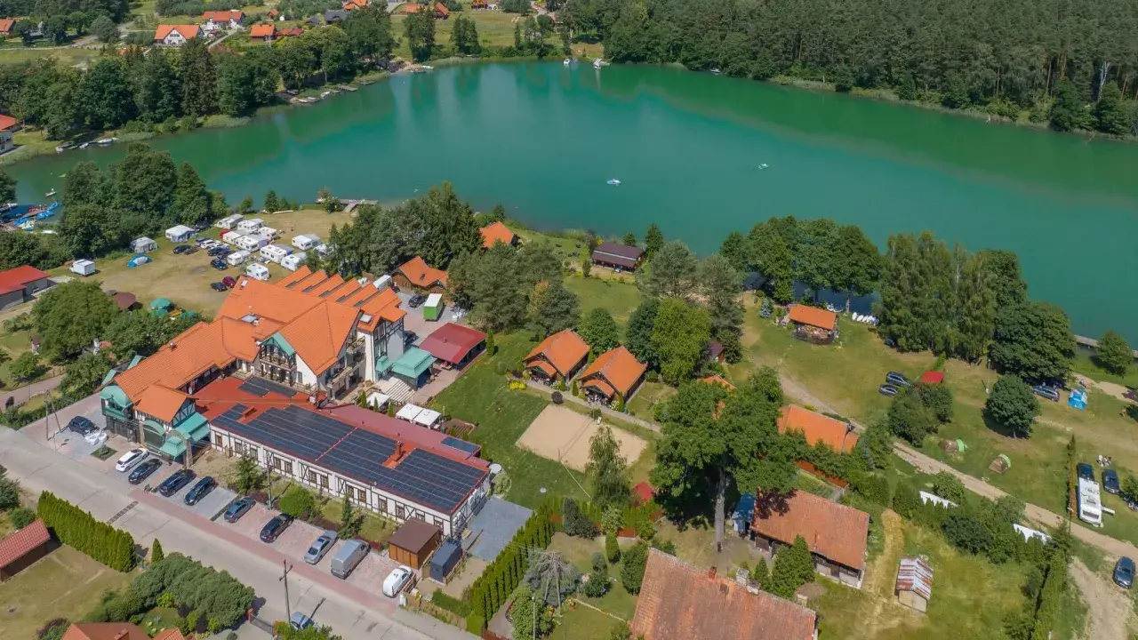 Widok z lotu ptaka na ośrodek wypoczynkowy nad turkusowym jeziorem. Widać budynki, domki, pole namiotowe, kampery i plażę. To idealne miejsce na wypoczynek, niczym złota łania wiartel.