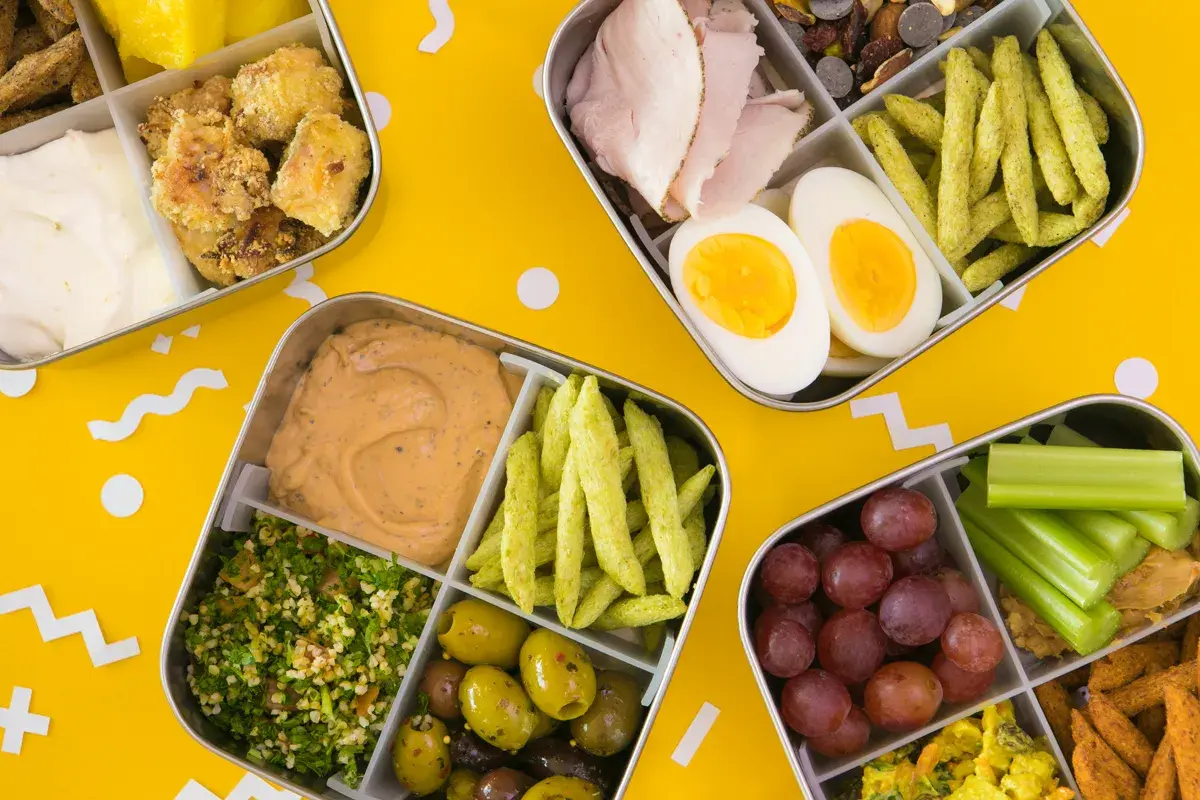 Four bento boxes filled with colorful, healthy foods, perfect for bento for beginners. Includes chicken, eggs, olives, grapes, and more.