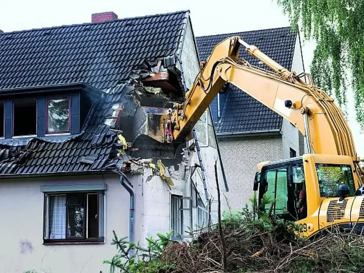 Żółta koparka prowadzi rozbiórkę dachu domu, niszcząc jego konstrukcję.