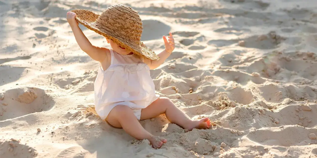 Liten bebis på stranden med solskyddsfaktor, leker med en stor stråhatt.