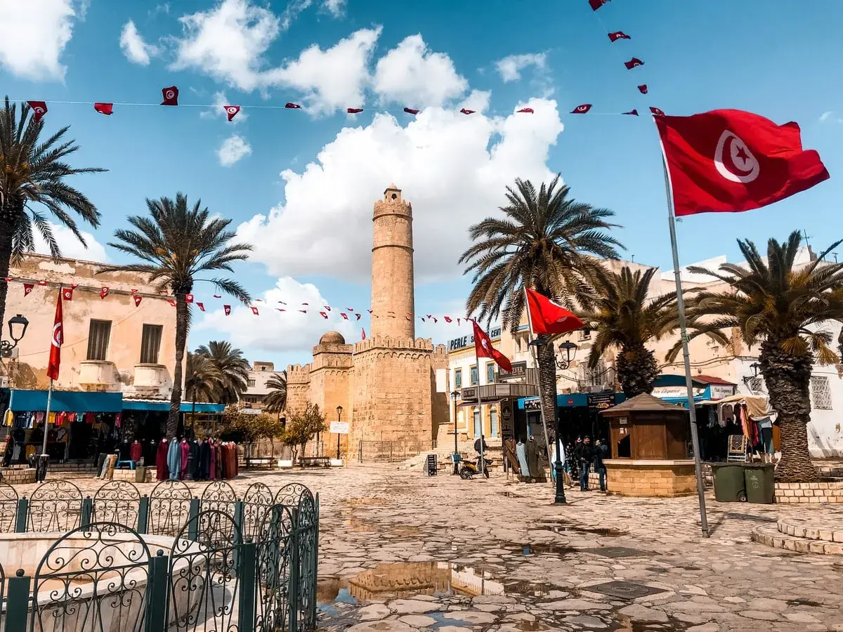 Placez-vous ici pour savoir où aller en Tunisie : une place animée avec des drapeaux tunisiens, des palmiers et une tour historique.