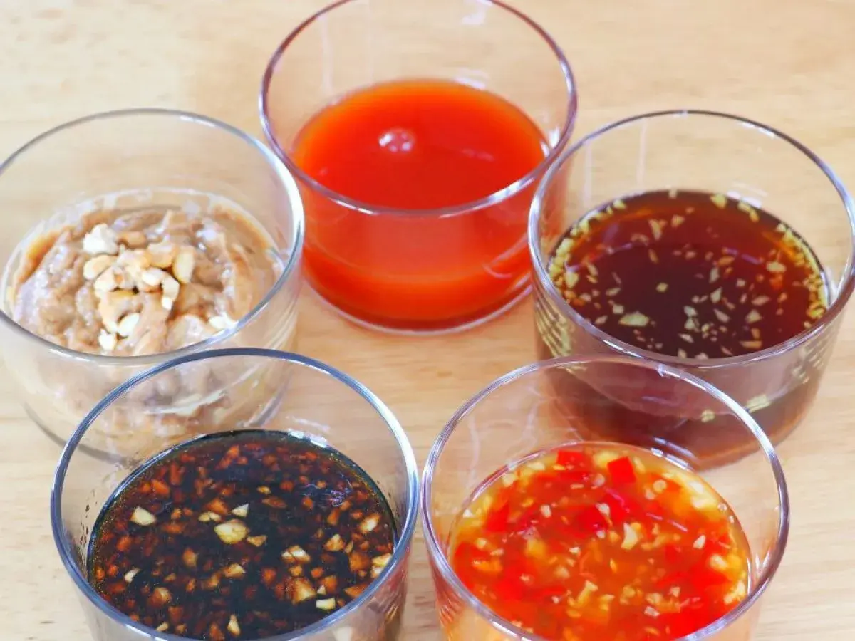 Five small glasses hold various Japanese dipping sauces, including a creamy peanut sauce, a bright red sauce, and a dark soy-based sauce with garlic.