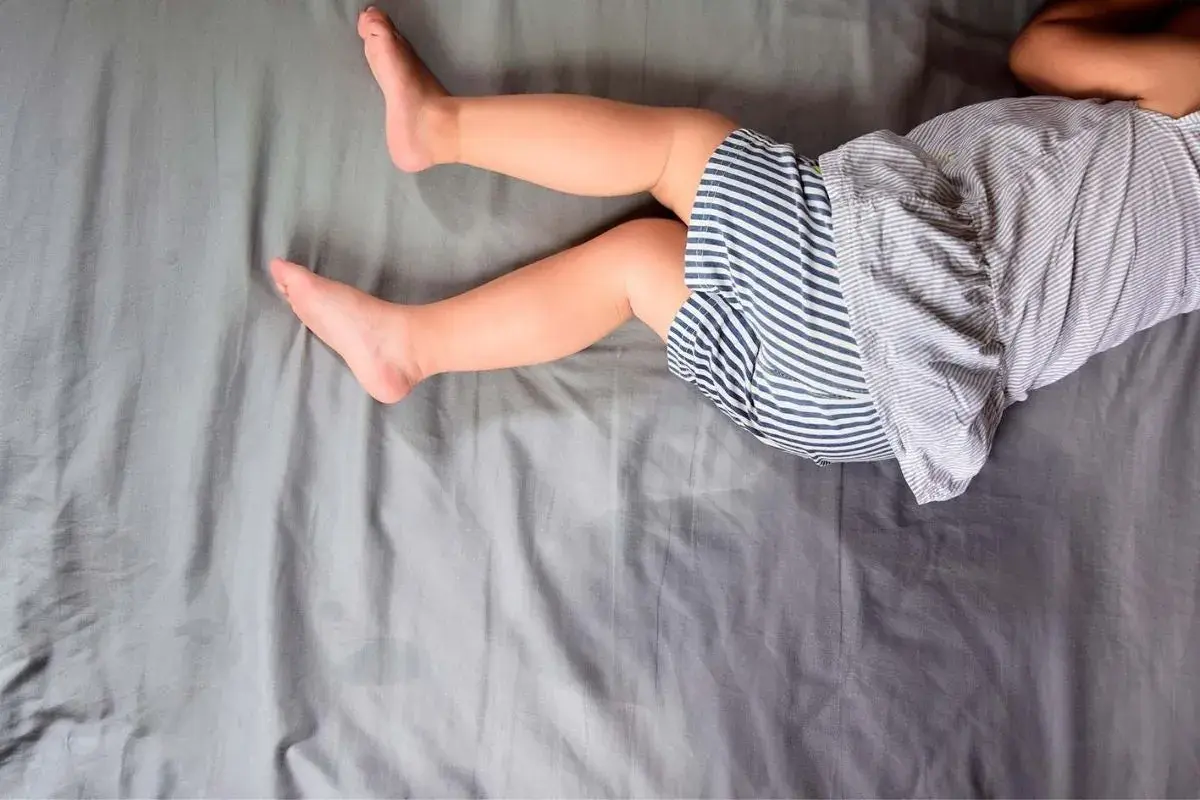 Gambe di bambino su lenzuola grigie. Un piccolo incidente notturno, forse legato all'enuresi notturna, ha lasciato un segno.