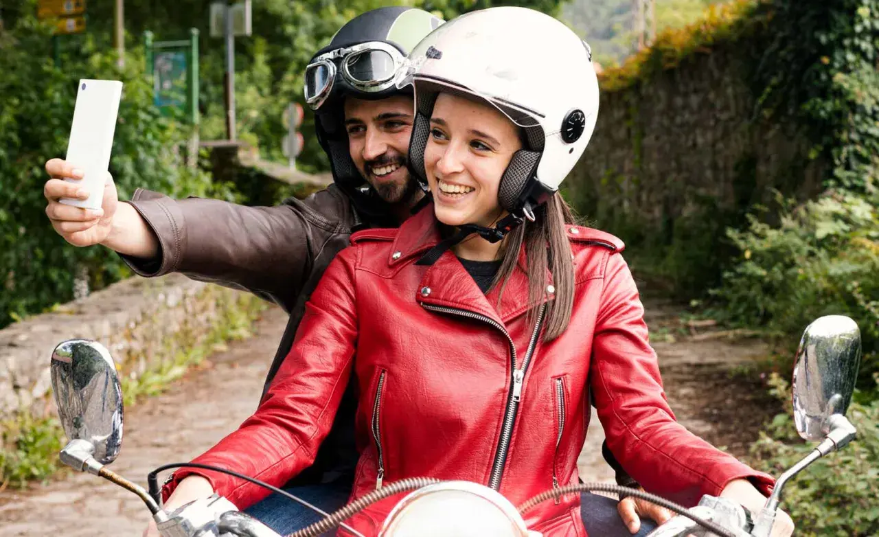 Pareja en moto, él con casco y gafas, ella con casco blanco y chaqueta roja, toman una selfie con el móvil. ¡Manos libres, casco moto legal y a disfrutar del viaje!
