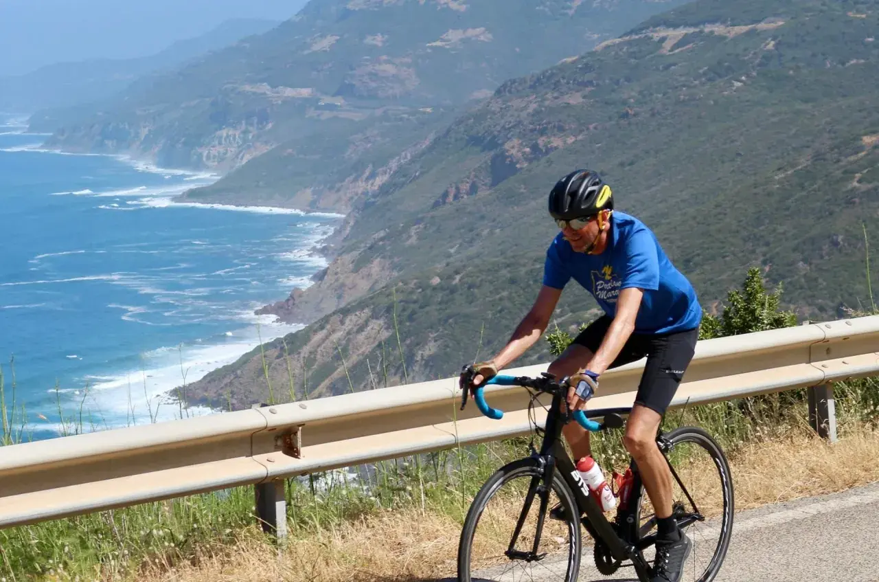 Man on a bike tour in Sardinia, cycling along a scenic coastal road with the blue sea and green hills.