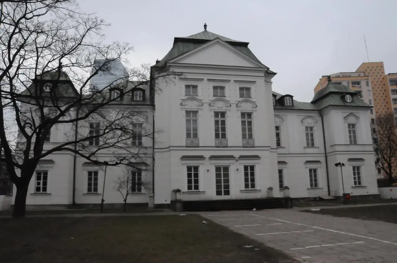 Pałac Przebendowskich, biała, elegancka budowla z zielonym dachem, otoczona drzewami i nowoczesnymi budynkami.