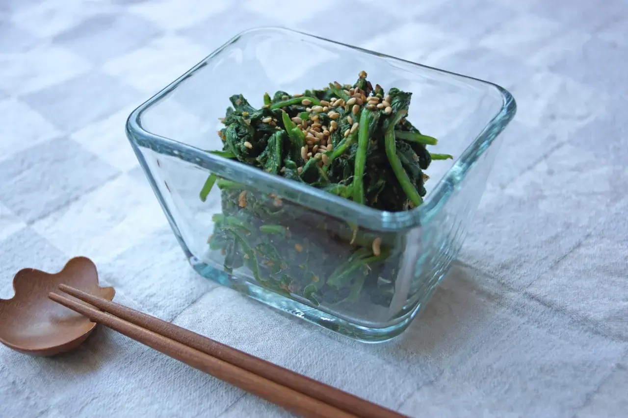 A glass bowl filled with spinach gomae, a savory sesame-dressed spinach dish, is ready to be enjoyed with chopsticks.