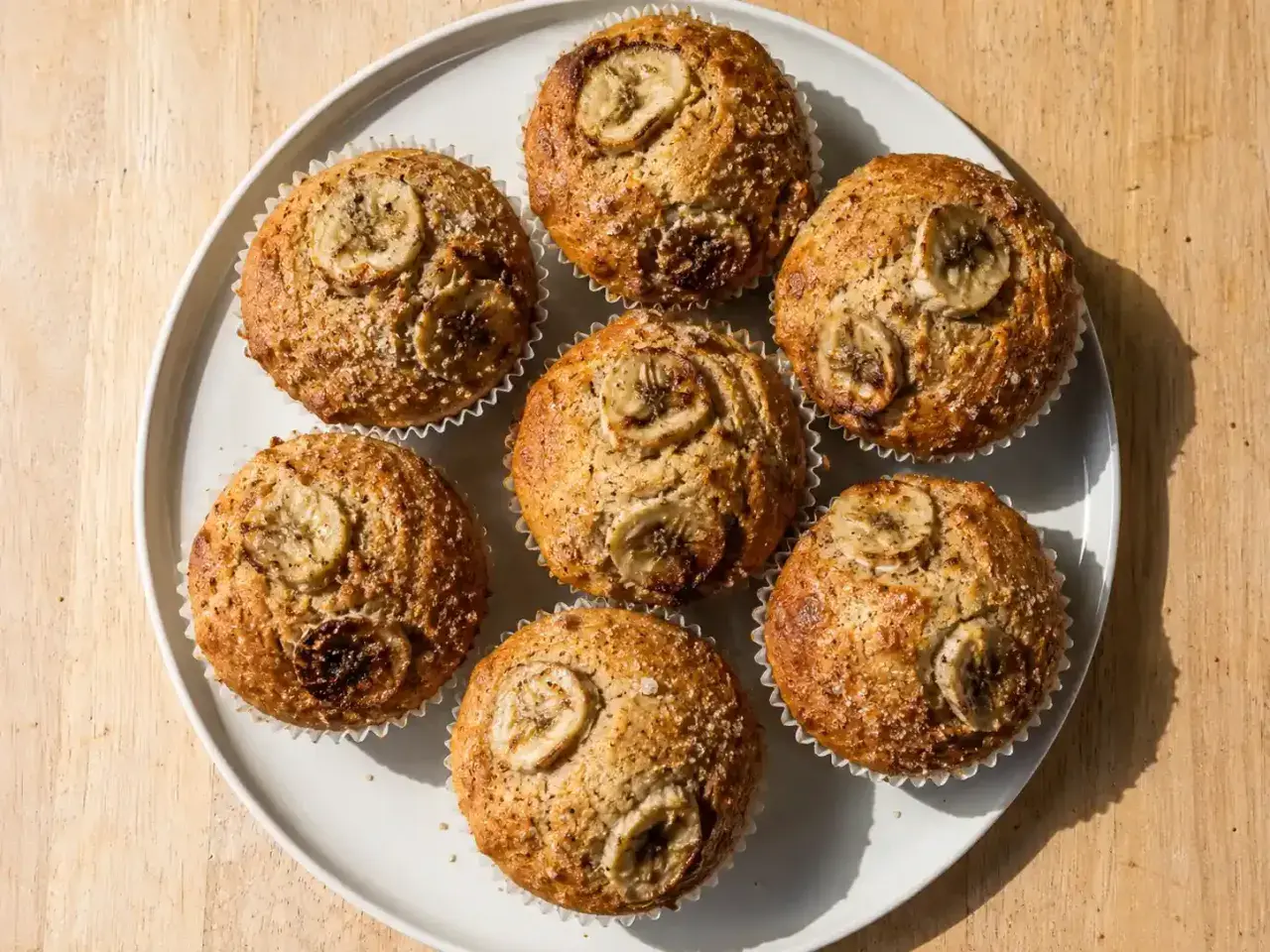 Sju gyllene bananmuffins med skivor av banan på toppen, serverade på en vit tallrik. Perfekt för fika!