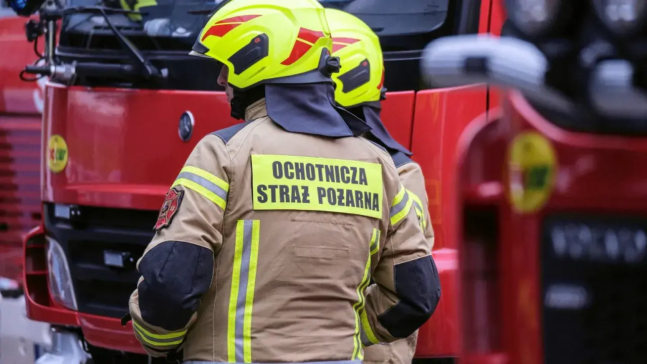Strażacy w żółtych hełmach i beżowych kurtkach z napisem "Ochotnicza Straż Pożarna" stoją obok czerwonego wozu strażackiego.
